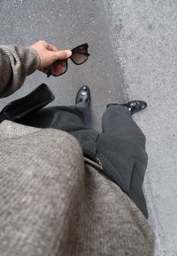 Melni saulesbrilles tur rokā, mugurā teksturēta pelēka džemperis un pelēki pārbaudītas bikses. Melni apavi redzami uz bruģētas virsmas.