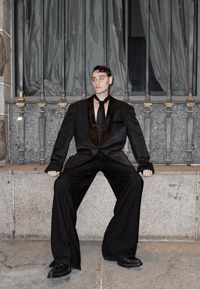 Traje negro con acentos de satén, pantalones de gran tamaño y una corbata a juego. Zapatos brillantes. Fondo gris con barras de metal y tela drapeada.