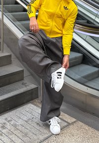 Sárga cipzáras dzseki fekete díszítéssel, szürke bőszárú nadrág és fekete csíkos fehér sportcipő, lépcsőn elhelyezve, mozgólépcső mellett.