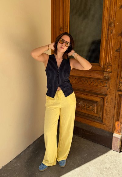 Chaleco a rayas azul marino con cuello en V, pantalones amarillos de pierna ancha y zapatos azules. Fondo de paneles de madera con detalles ornamentales.