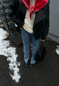 Personne portant une veste en cuir noire, un pull crème, une écharpe rouge, un jean bleu et des chaussures noires, tenant un sac à main noir sur un sol pavé enneigé près des escaliers.