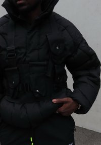Fekete, szigetelt puffer dzseki zsebekkel és pántos részletekkel, magas gallérral és behúzott aljjal. Az anyaga fényesnek és párnázottnak tűnik.