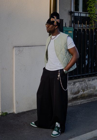 Chaleco acolchado verde claro sobre una camiseta blanca, pantalones anchos negros y zapatillas verdes. Gorro negro con cruz decorativa y acentos de cadena plateada.