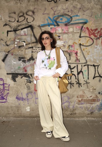 Camisa blanca de manga larga con estampado floral colorido, pantalones de pierna ancha a rayas beige, zapatillas negras y blancas, y un bolso de hombro marrón.