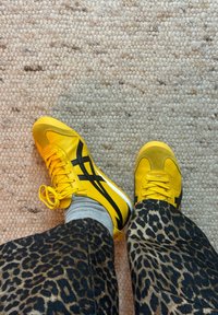 Amarillos zapatos deportivos con detalles negros, materiales de cuero y ante, cordones estándar y suelas de goma blanca. Pantalones con patrones al fondo.