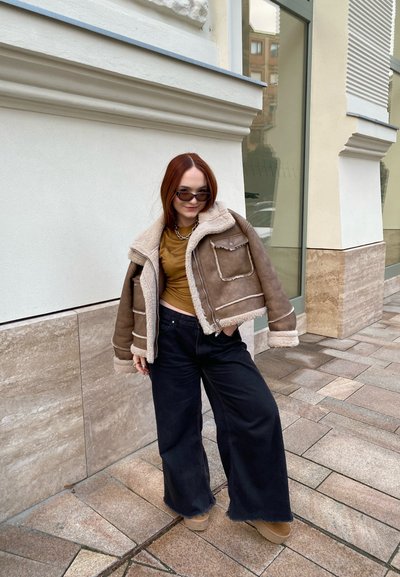 Chaqueta recortada de borreguillo marrón con acentos beiges, combinada con una blusa marrón y pantalones amplios negros. Estilizada con gafas de sol oscuras y botas beiges.