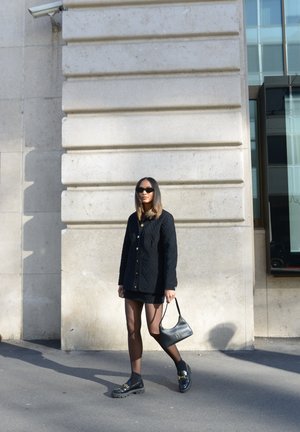 Mujer con chaqueta acolchada negra, falda corta, medias transparentes, mocasines negros, gafas de sol, sosteniendo un pequeño bolso negro mientras camina por la acera de la ciudad.