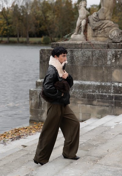 Persona con una chaqueta negra con cuello de piel y pantalones anchos oliva subiendo unas escaleras de piedra junto a un lago, con una estatua de piedra tallada cerca.