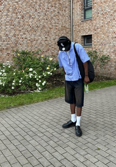 Gorra negra, camisa de manga corta azul, pantalones cortos de mezclilla, calcetines blancos, zapatos negros, sosteniendo una bebida, de pie en un pavimento cerca de flores.