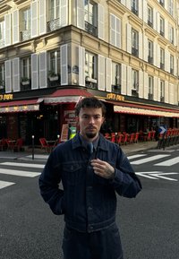 Jeansjacka och byxor i mörkblått, med fickor och en figurnära design. Randig skjorta under, med en slips och en tatuerad hand synlig.