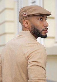 Hnědý plochý klobouk s logem Lacoste, nošený osobou v béžovém tričku s dlouhým rukávem. Detailní záběr ukazuje texturu a designové detaily.