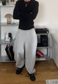 Persona in maglietta nera a maniche lunghe e pantaloni grigi con le braccia incrociate accanto a uno scaffale metallico che tiene un giradischi, un altoparlante e dischi.