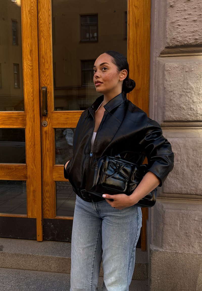 Femme aux cheveux foncés attachés en chignon, portant une veste en cuir noire et un jean bleu clair, tenant un sac à main noir, debout près de portes en bois et d'un mur en pierre.