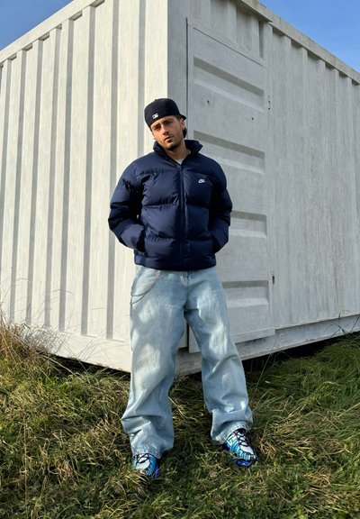 Chaqueta de plumas azul navy, jeans sueltos de lavado claro y zapatillas coloridas. De fondo se observa un contenedor de almacenamiento blanco y césped verde.