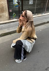 Mladá žena s dlouhými vlasy, slunečními brýlemi, béžovým šátkem s puntíky, pískovou bundou a robustními teniskami dřepí na městském chodníku poblíž zavřeného obchodu.