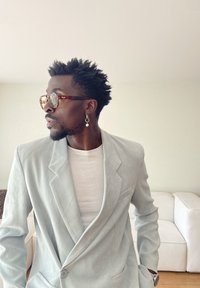 Muž s krátkymi kučeravými vlasmi, ktorý má na sebe korytnačkové slnečné okuliare, perlovú náušnicu, svetlosivý blejzer a bielu košeľu, stojí v interiéri vedľa bielej pohovky.