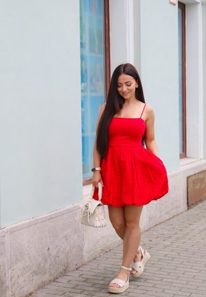 Vestido rojo sin mangas con falda acampanada, llevado con sandalias beige y un bolso blanco. Pelo largo y liso con un fondo claro.