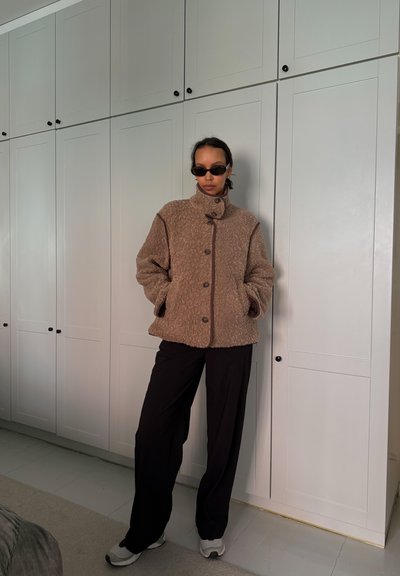 Persona que lleva una chaqueta de forro polar marrón con botones, pantalones anchos negros, zapatillas deportivas blancas y gafas de sol negras, de pie frente a puertas de gabinete blancas.