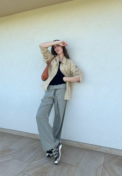 Camisa overshirt beige, camiseta negra, pantalones holgados verde olivo y zapatillas blanco y negro. Bolso tejido marrón y pared texturizada de fondo.
