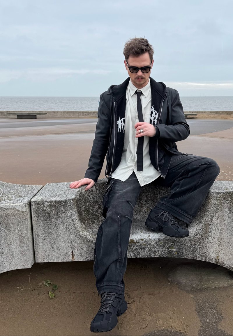 Mann iført svart jakke, hvit skjorte, svart slips og solbriller sitter på en buet betongbenk ved sjøen på en overskyet dag.