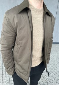 Mann iført olivengrønn jakke med glidelås over beige strikkegenser og svarte bukser, stående på flislagt fortau med grå vegg i bakgrunnen.