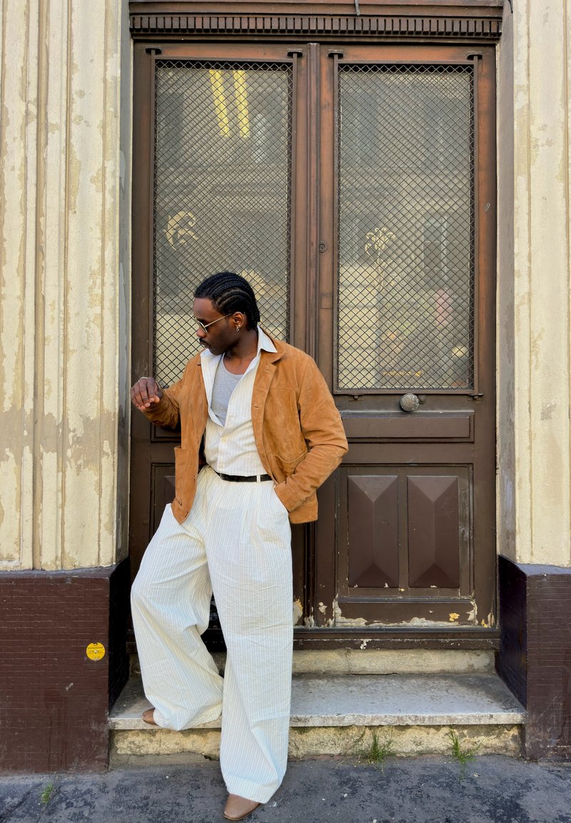Homme aux cheveux tressés portant des lunettes de soleil, une veste brune et un pantalon blanc large, debout sur une marche devant une grande porte brune avec un grillage en fil de fer.