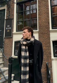 Ένας νέος άνδρας φοράει ένα μαύρο παλτό και μια καρό μαύρη και μπεζ φου scarf, στέκεται δίπλα σε έναν τοίχο από τούβλα και σε μια ταμπέλα που γράφει "DE VLIEG."