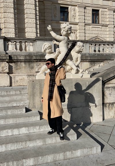 Abrigo trench beige, bufanda negra con patrón de cuadros, pantalones negros y zapatillas. Escaleras de piedra y esculturas blancas al fondo.