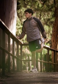 Mees spordivarustuses sirutab ühte jalga, hoides käega piirdetõkke äärest metsasees asuval teel päeval.