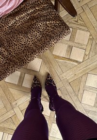 Talons pointus à imprimé léopard portés avec des leggings violets, debout sur un parquet en bois. Un siège en peluche à imprimé léopard est à proximité.