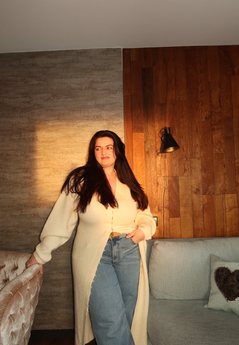 Femme aux longs cheveux foncés portant un cardigan crème et un jean, debout à côté d'un fauteuil en velours dans un coin chaleureux et doucement éclairé d'un salon.