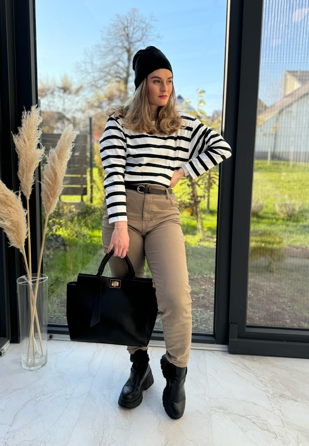 Μαύρη και λευκή ριγέ μακρυμάνικη μπλούζα, μπεζ παντελόνι με ψηλή μέση, μαύρες μποτάκια μέχρι τον αστράγαλο και μια μεγάλη μαύρη τσάντα. Φυτά στο φ�όντο.