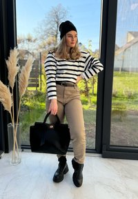 Μαύρη και λευκή ριγέ μακρυμάνικη μπλούζα, μπεζ παντελόνι με ψηλή μέση, μαύρες μποτάκια μέχρι τον αστράγαλο και μια μεγάλη μαύρη τσάντα. Φυτά στο φόντο.