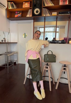 Suéter crema de gran tamaño con lazo rosa, falda de cuero verde, calcetas rosa claro, tenis amarillos y un bolso verde sobre un taburete.