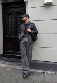 Černá kožená bunda, černé tričko, světlé šedé široké džíny, černé tenisky a černá baseballová kšiltovka. Tmavé dřevěné dveře v pozadí.