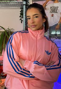 Jeune femme aux cheveux foncés, portant des boucles d'oreilles anneaux et un anneau au nez, vêtue d'une veste de survêtement rose Adidas avec des bandes bleues, les bras croisés dans un cadre intérieur décontracté.