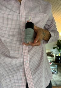 Szary antyperspirant w formie sticka o cylindrycznym kształcie z czarną nakrętką; trzymany przed w kratkę różowo-białą koszulą z guziczkami.