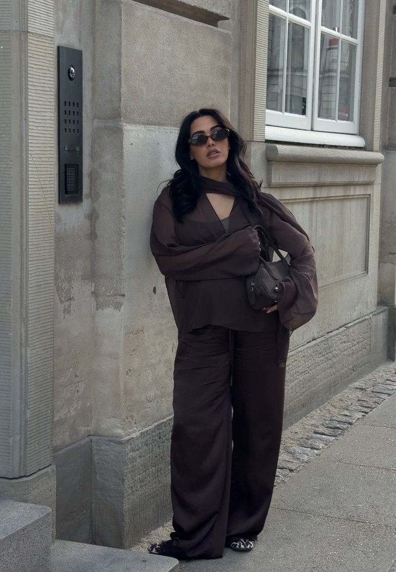 Woman wearing dark loose clothing and sunglasses stands against stone building wall holding a small bag on a city street.
