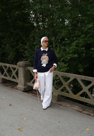 Marineblauer Pullover mit Bärenmotiv, darunter ein weißes Hemd mit Kragen, weite weiße Hose und eine rosa Handtasche; kombiniert mit einem weißen Kopftuch.