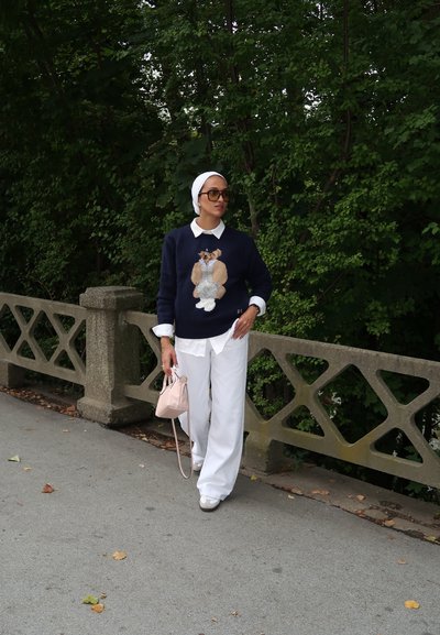 Suéter navy con gráfico de oso, camisa de collar blanco debajo, pantalones amplios blancos y bolso rosa; estilizado con un pañuelo blanco en la cabeza.