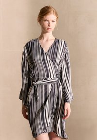 Vestito avvolgente con maniche lunghe a righe bianche e nere, dotato di una cintura in vita e di un tessuto morbido e liscio. I bordi esterni presentano una leggera ondulazione.