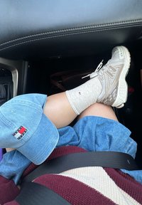 Sneaker New Balance grigi, calze grigie chiare, un cappellino in denim blu e una gonna blu, con un maglione a strisce bordeaux e crema e una cintura di sicurezza nera.