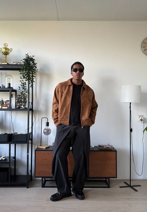 Hombre con gafas de sol, chaqueta marrón y pantalones negros anchos está de pie en una sala de estar moderna con estanterías, lámpara y reloj en la pared.