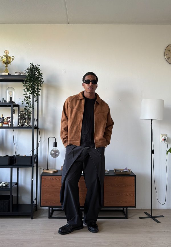 Mężczyzna w okularach przeciwsłonecznych, brązowej kurtce i szerokich czarnych spodniach stoi w nowoczesnym salonie z półkami, lampą i zegarem na ścianie.