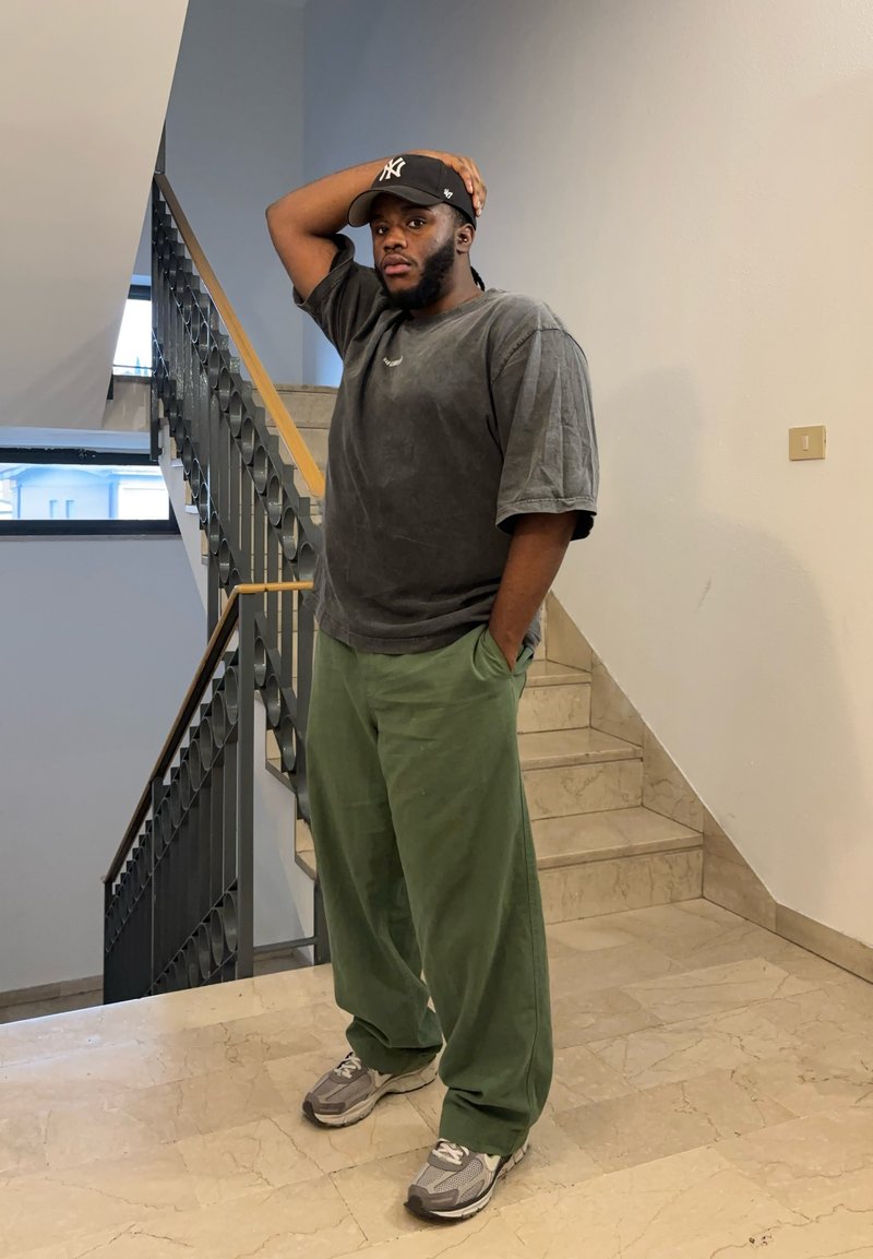 Gray oversized T-shirt with short sleeves, green loose-fitting pants, and gray sneakers. Standing on a staircase with a hand on the head.