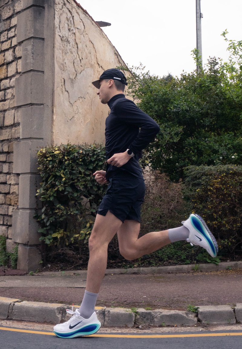 Man wearing black cap, long-sleeve shirt, and shorts running on sidewalk beside stone wall and greenery.