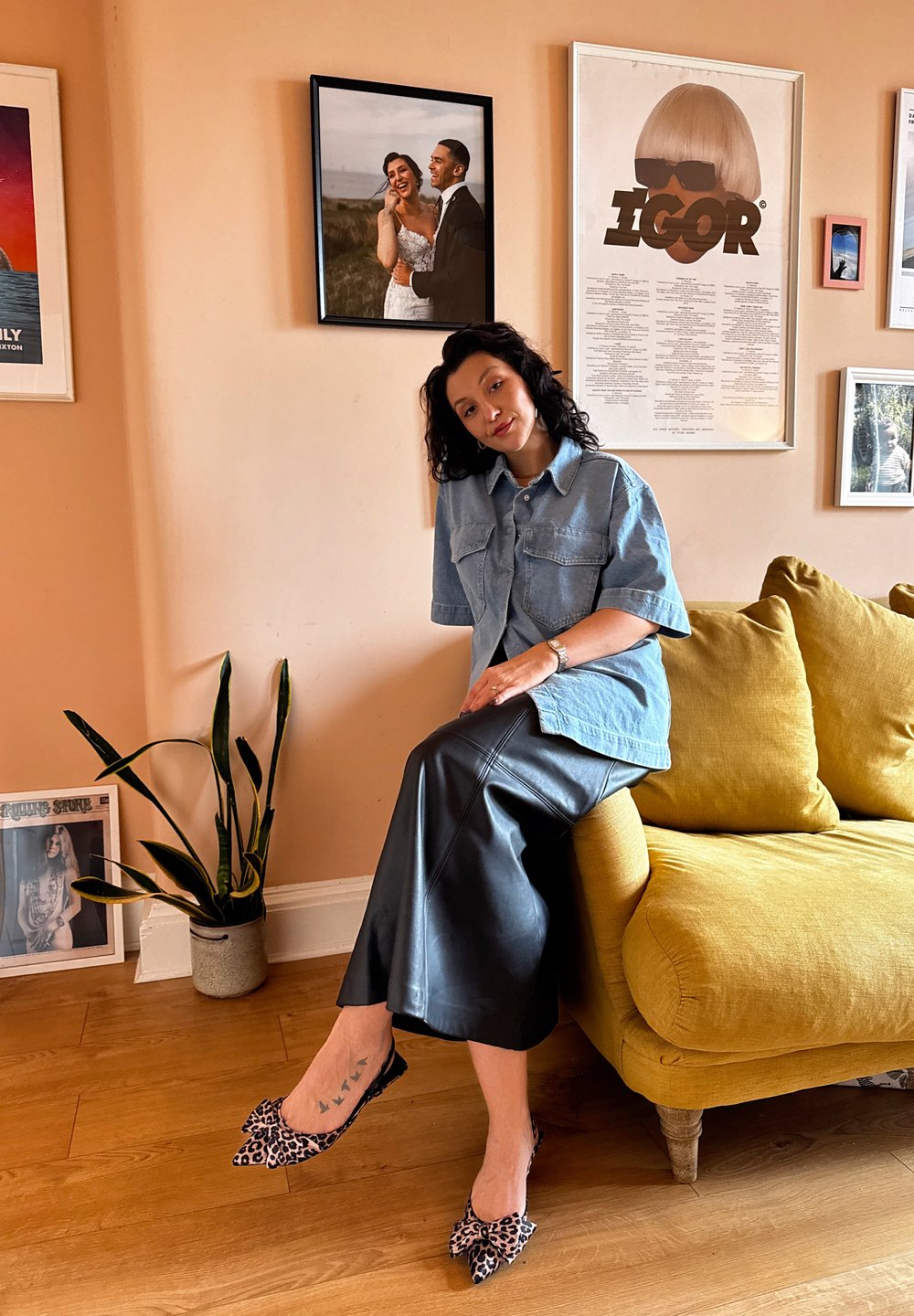 Camisa de manga curta em denim, saia de couro preta e sapatilhas com padrão animal. Sofá amarelo e planta numa sala de tons quentes com arte emoldurada.