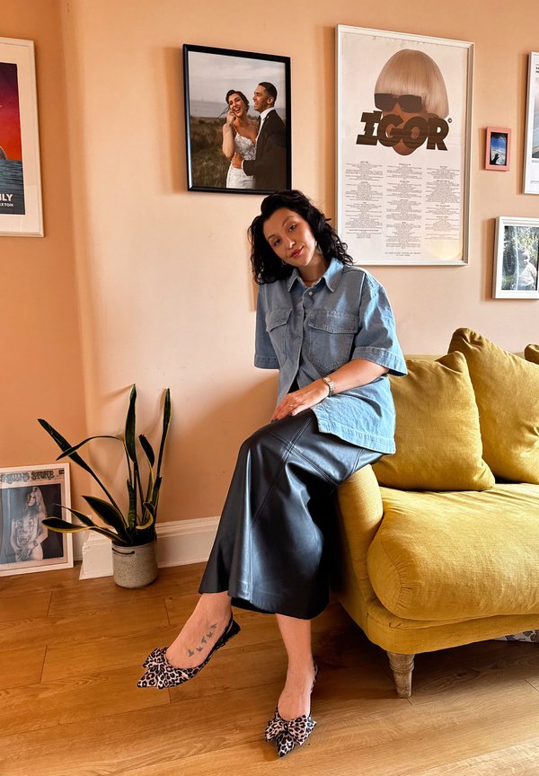 Denim kortærmet skjorte, sort lædernederdel og dyreprint flade sko. Gul sofa og plante i et rum med varme nuancer og indrammet kunst.