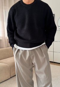 Pull noir en maille côtelée avec un col rond, porté sur un t-shirt blanc, associé à un pantalon plissé beige. Tissu texturé et coupe décontractée.