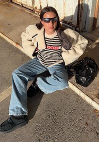 Jeune femme portant des lunettes de soleil, un pull rayé avec l'inscription "ADORE", une veste en peau retournée crème, un jean bleu clair et des chaussures noires, assise sur le trottoir près d'un sac noir.